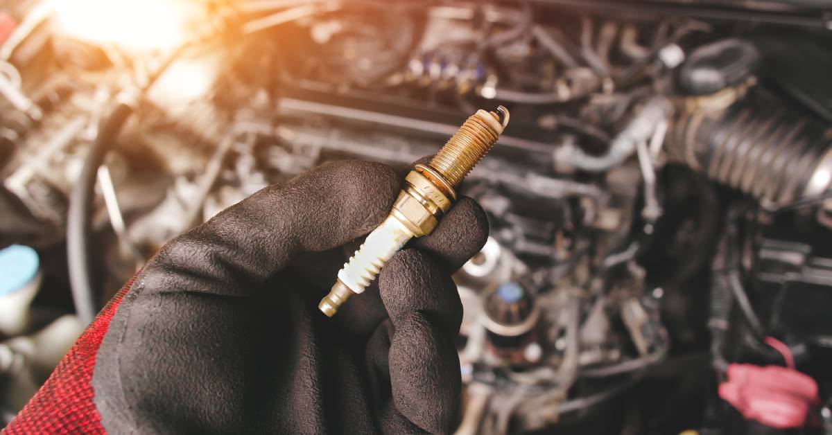 A gloved hand holding an old iridium automotive spark plug with the exposed engine in the background.