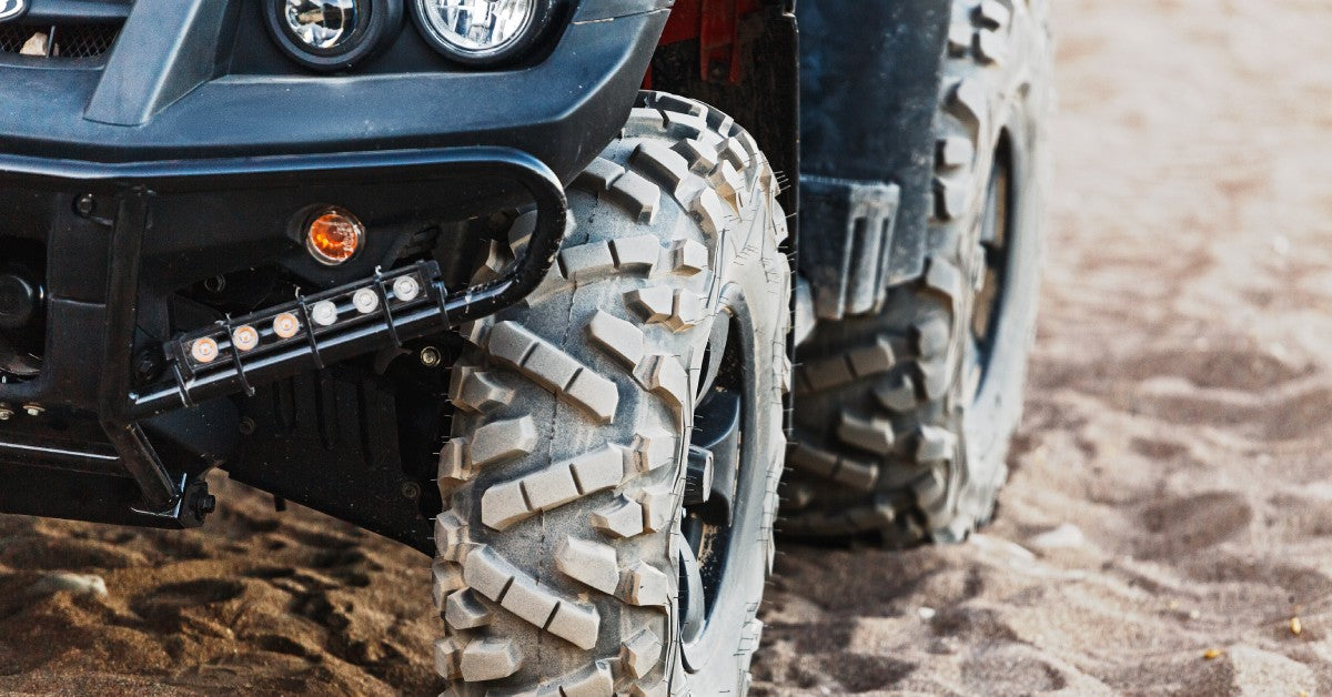 The front and rear tires of an all-terrain vehicle sit on sandy terrain. The ATV has off-road tires.