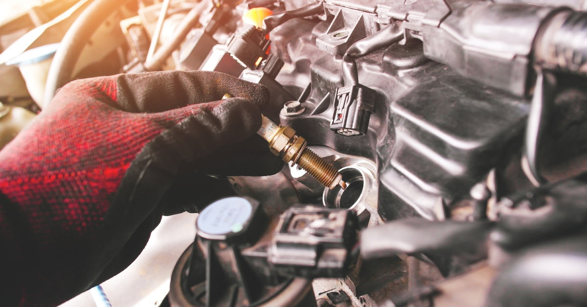 A mechanic wearing a red glove holds a copper spark plug near the ignition socket of a vehicle's engine compartment.