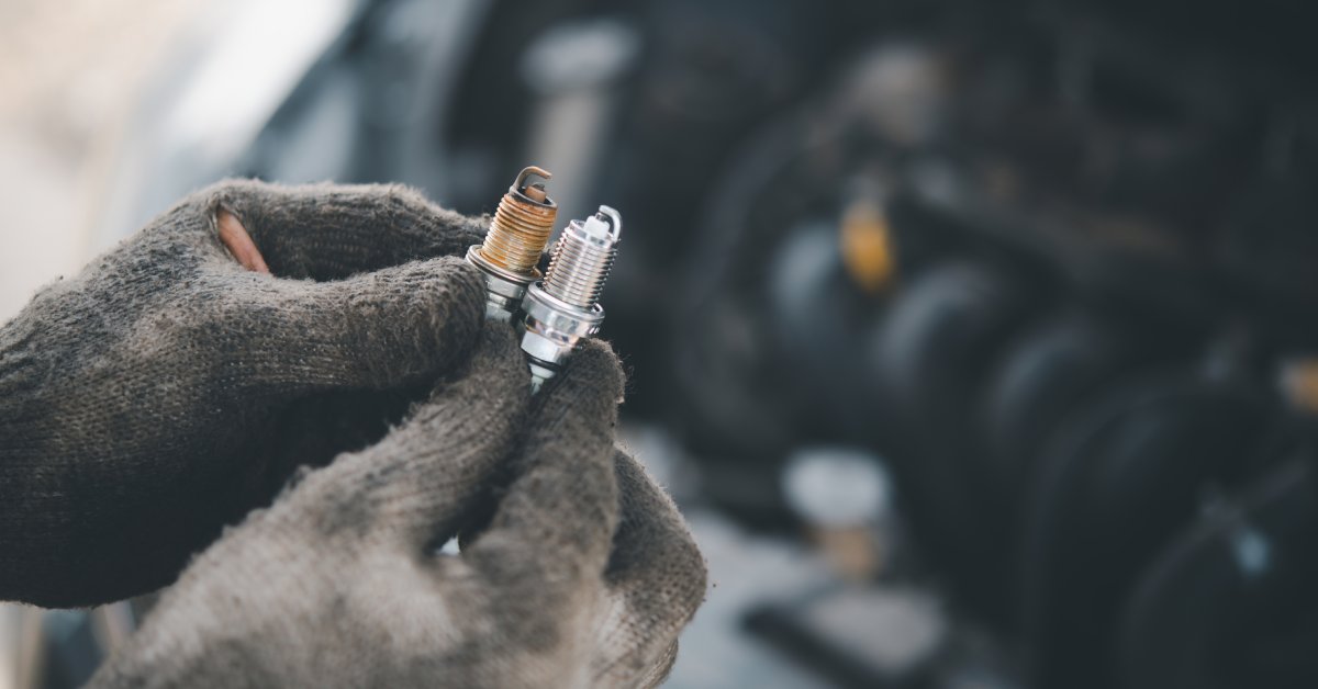 A pair of gloves, hands in focus, are holding an old and new spark plug together. An engine is blurred in the background.