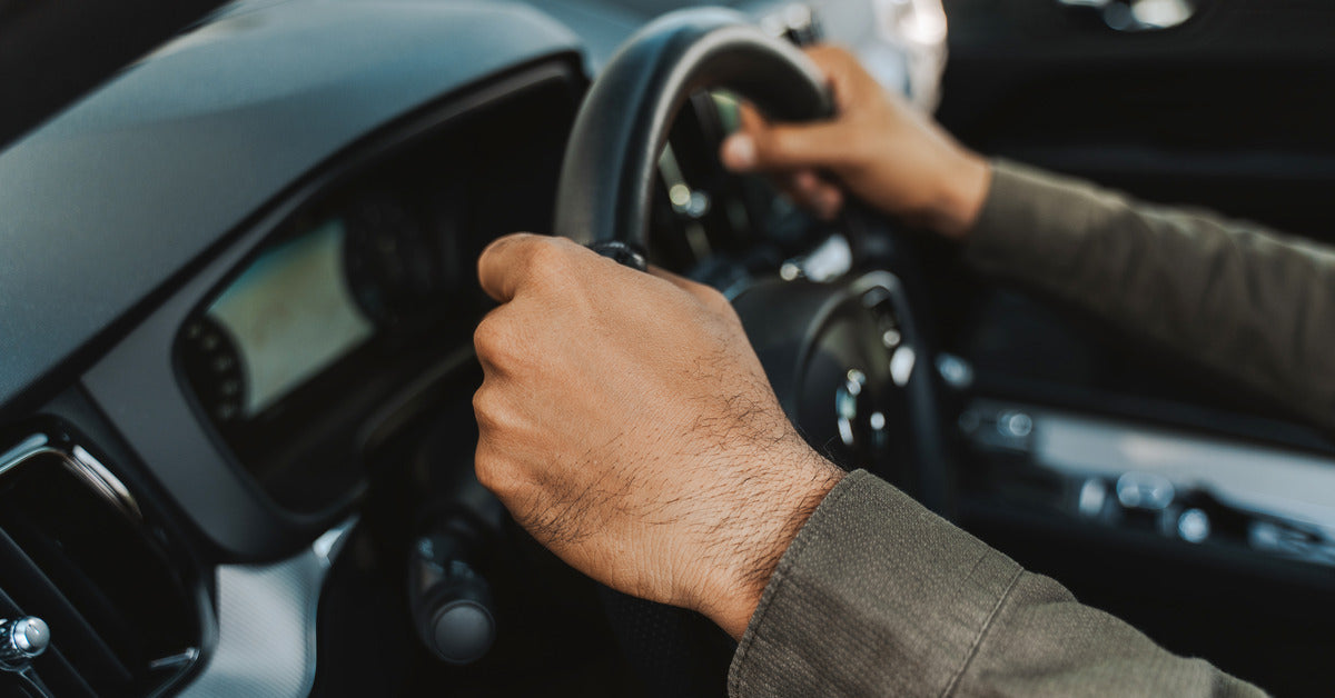 A close-up of a man with long sleeves holding a car steering wheel. The focus is on his hands gripping the wheel.