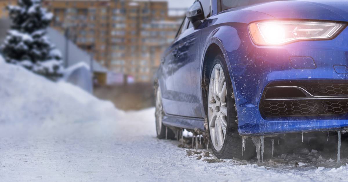 A blue car with the headlight on. There are icicles hanging from the bottom of the car and snowy conditions in the environment.