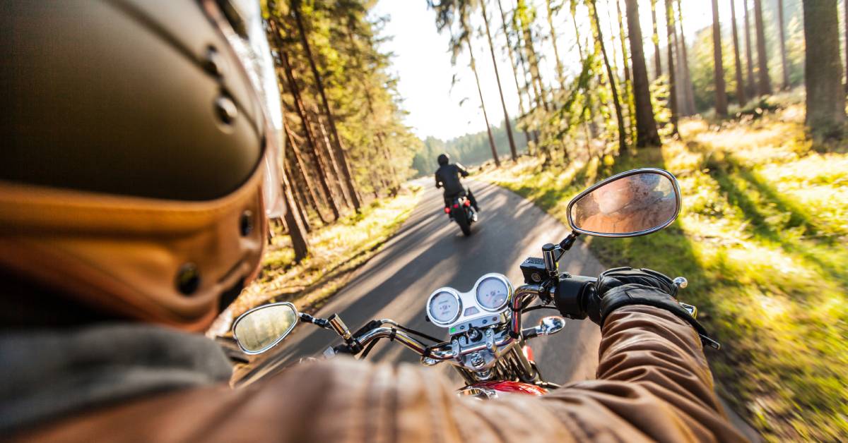 An over-the-shoulder view of a motorcycle rider following another motorcycle rider through a forest road.