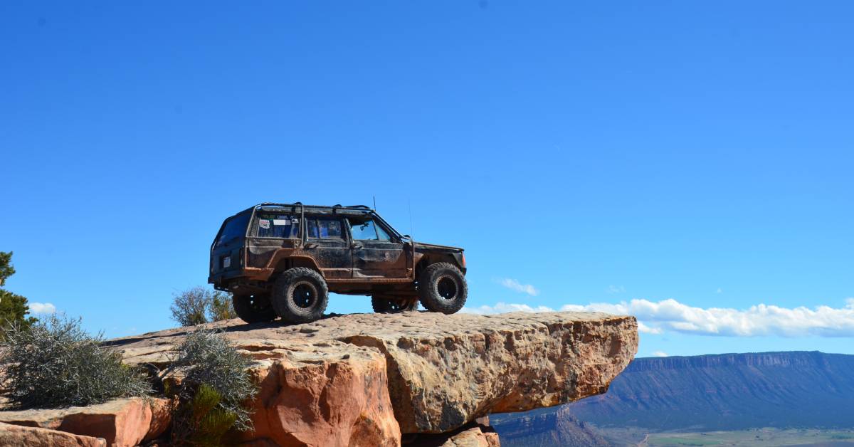 An off-road truck sitting at the edge of a cliff, looking out into an open landscape. It is a sunny day with minimal clouds.