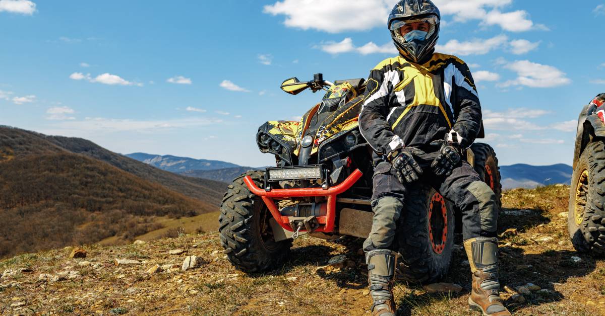 In a mountainous environment, a man wearing professional gear and a helmet sits on one of the tires of the ATV beside him.