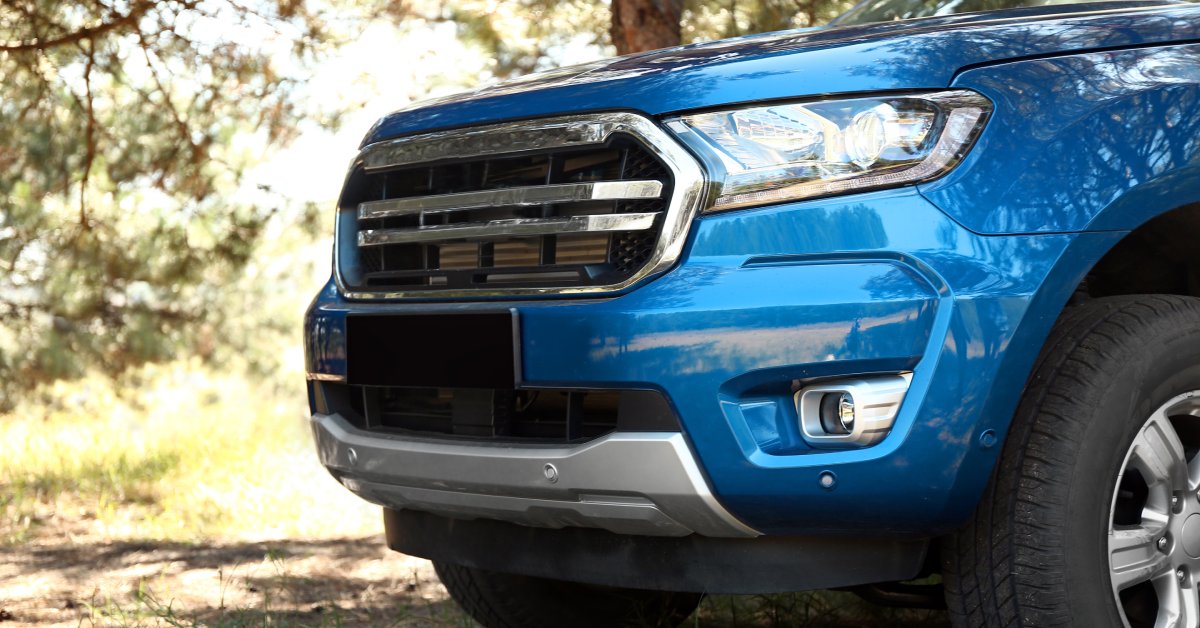 A blue pickup truck is parked under the shade of some trees on a sunny day, in what appears to be a countryside.