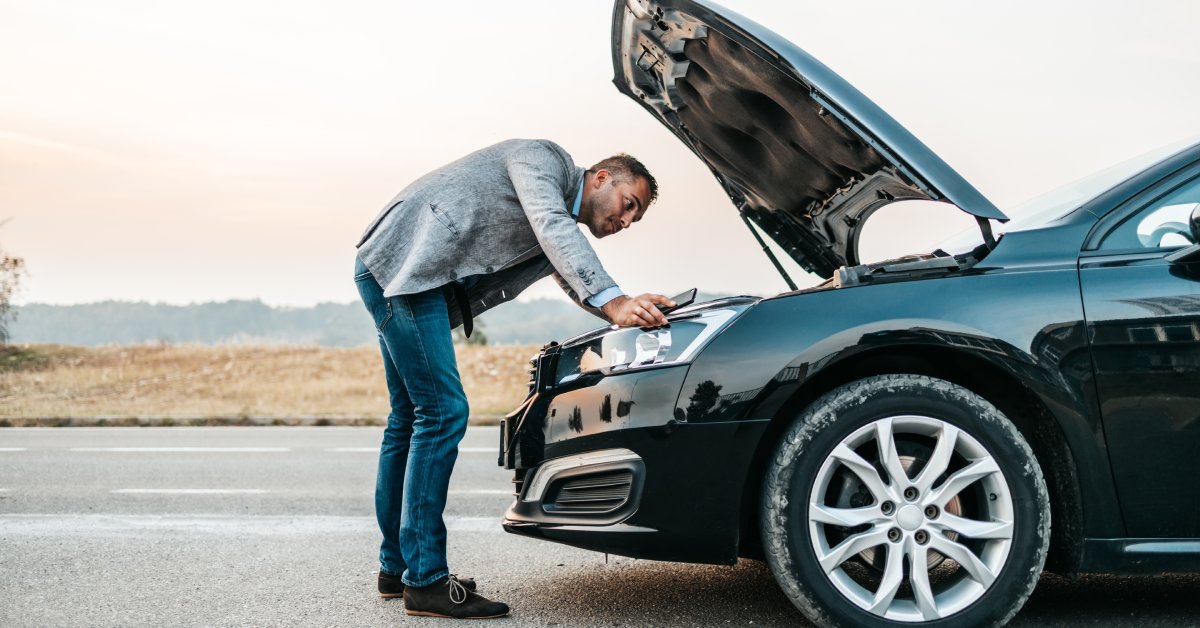 A well dressed man standing in front of his car's open hood. There is an open field and trees in the background.